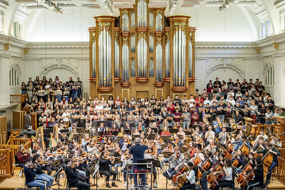 A male conductor rehearsing an orchestra performance, with a orchestra surrounding him performing, with a group of singers singing in the back, with the organ behind them.